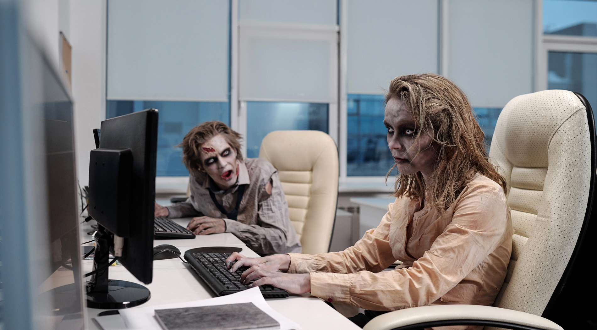 Two office workers perform routine tasks in a modern workplace, representing an organization in zombie mode — active in form, but lifeless in identity, collaboration, and creative renewal.