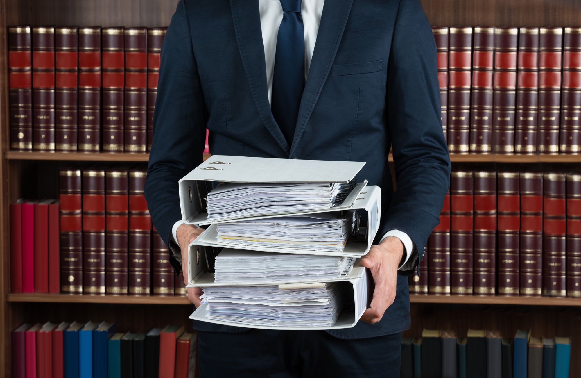 A suited professional holding stacked binders full of legal documents in front of a law library.